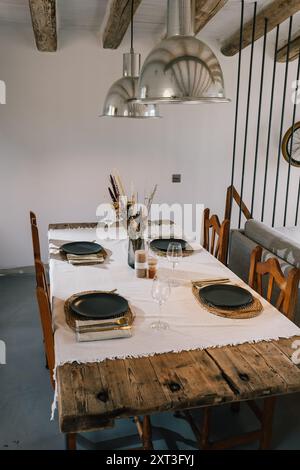 Beautifully arranged dinner table in a rustic dining room featuring a vintage wooden table, golden black plates on wicker placemats, and chic glasswar Stock Photo