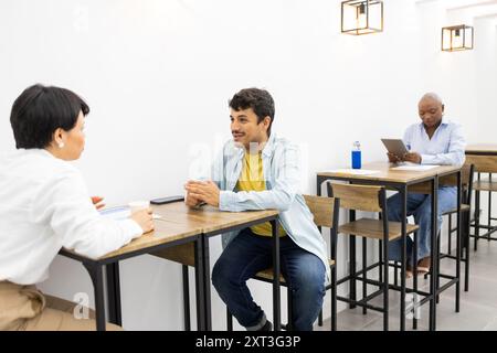 A vibrant English academy scene featuring adults in a modern classroom setting, engaging in dialogue across wooden desks while a fellow student works Stock Photo
