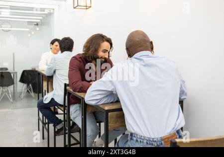 In a bright, modern English academy, two students engage in a focused discussion while sitting at a high table Other participants converse in the back Stock Photo