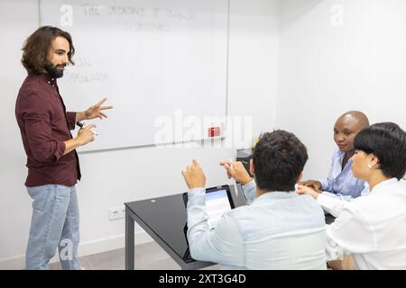 In a brightly lit classroom, a male teacher gestures animatedly while explaining concepts to a diverse group of attentive adult students, highlighting Stock Photo
