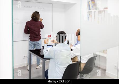 An English language class shows students and a teacher interacting The teacher writes on a whiteboard while discussing with students seated around a t Stock Photo