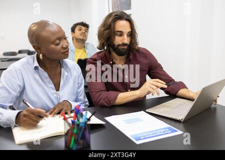In a modern classroom setting, a diverse group of three professionals collaborates over a laptop discussing academic work, highlighting teamwork in an Stock Photo