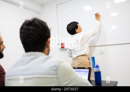 A professional woman is actively teaching in a modern classroom with attentive students She writes on a whiteboard, facilitating an engaging English l Stock Photo