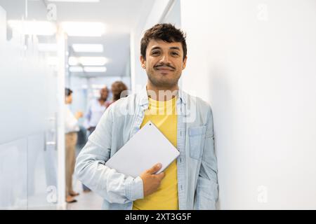 A cheerful young man holding a digital tablet stands in a brightly lit educational hallway, smiling with a background of peers discussing Stock Photo