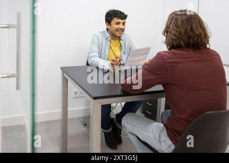 Two individuals engaged in a discussion at a desk within a bright, modern classroom environment, exemplifying effective collaboration and teamwork in Stock Photo