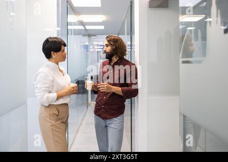 A man and woman converse while holding coffee mugs in a sleekly designed corporate hallway, illustrating a casual yet focused work discussion Stock Photo