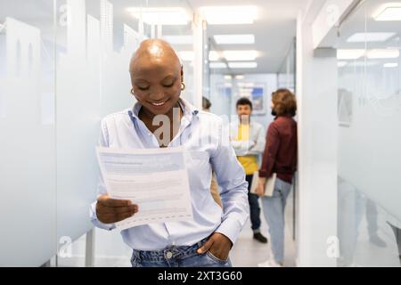 A young woman reviews papers in a bustling academy corridor, emphasizing teamwork in an English learning environment Colleagues engage in discussions Stock Photo