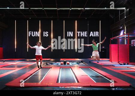 A cheerful young man and woman mid-air at a vibrant trampoline park, surrounded by colorful equipment, under a sign stating FUN IS IN THE AIR. Both lo Stock Photo
