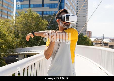 Middle-aged Indian man in a yellow shirt uses a VR headset while stretching on a city bridge. Stock Photo