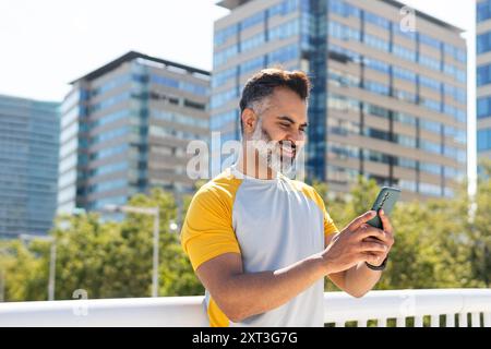 Middle-aged Indian man in a yellow and white sports shirt, smiling while using his smartphone outdoors. He is looking at the screen, standing with urb Stock Photo