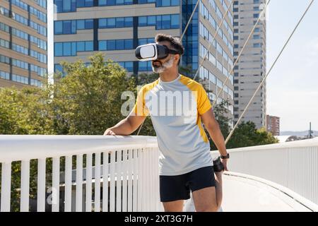 Indian man wearing a virtual reality headset stands on a city bridge, demonstrating the blend of technology and physical activity. He is dressed in sp Stock Photo