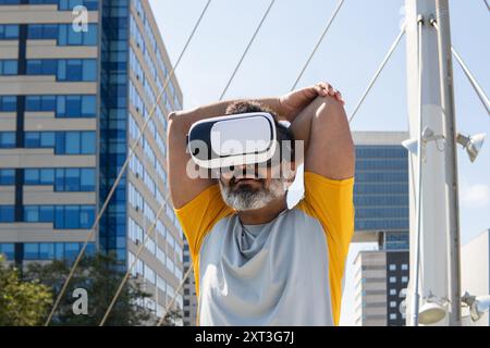 Indian man using virtual reality headset, experiencing technology outdoors with cityscape background. He is looking at screen, engaging with the immer Stock Photo