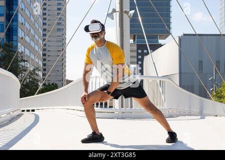 An Indian man is wearing a virtual reality headset and sports attire, stretching on a city bridge with skyscrapers in the background. He is looking do Stock Photo