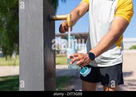 Cropped, unrecognizable thirsty man in sportswear filling a reusable water bottle at an outdoor faucet in a sunny park, showcasing a healthy, active l Stock Photo