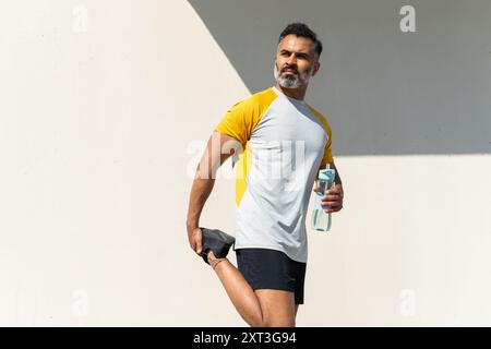 An Indian man looking away while stretching his leg and holding a water bottle during a summer workout outdoors. Stock Photo