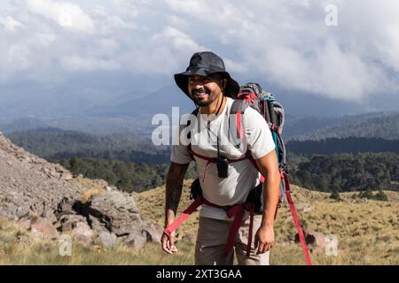 A cheerful male hiker with a beard and tattoos, wearing a sun hat, backpack, and equipped with trekking poles, looks directly at the camera with a wid Stock Photo
