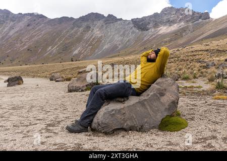 A man resting on a large rock with his arms behind his head, with eyes closed, amidst a rugged mountainous terrain. His expression exudes relaxation a Stock Photo