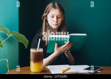 Female student sitting at a table, absorbed in reading a green book, with iced tea and a smartphone nearby. She is looking down, engaged in her study Stock Photo