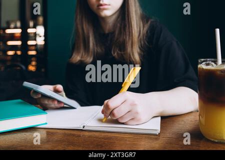 Cropped unrecognizable female student writing on her notebook with a yellow pen, with a smartphone in her other hand and a cold drink beside her on th Stock Photo