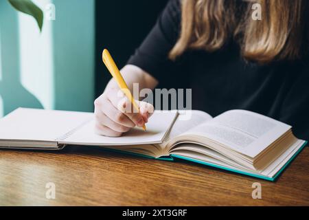 Cropped unrecognizable photo of a female student looking down while writing notes in a book, sitting at a wooden table, with a focus on her hands and Stock Photo