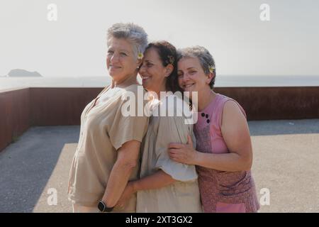 Three elderly female friends share a warm embrace by the seaside, feeling joyful. One women looks at the camera while the other two are looking down, Stock Photo