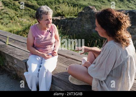 Two elderly deaf women engaging in a conversation using sign language while seated on a bench outdoors, exemplifying communication and friendship in o Stock Photo