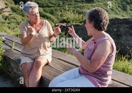 Two elderly deaf women engaging in a conversation using sign language while seated on a bench outdoors, exemplifying communication and friendship in o Stock Photo