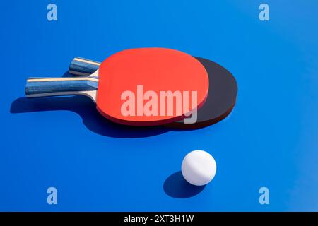 Vibrant image showing two table tennis paddles, one red and one black, along with a white ball, all set against a stark blue background Ideal for them Stock Photo