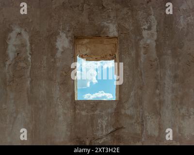 A decrepit window opening in an old, abandoned building frames a view of the serene blue sky with fluffy clouds. Stock Photo