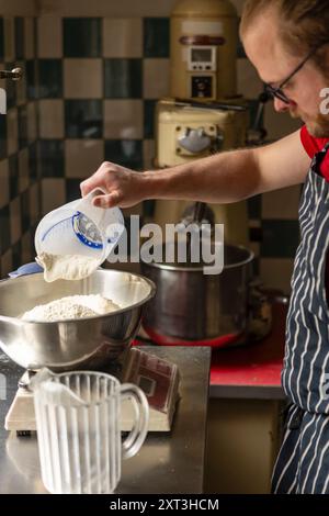 A focused man wearing glasses and a striped apron pours flour from a measuring cup into a large metal bowl in a vintage-style kitchen, preparing dough Stock Photo