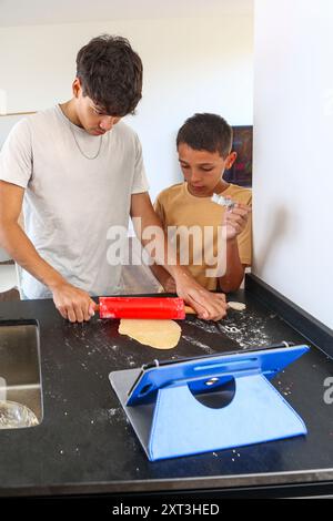 Siblings engage in baking, rolling out dough on a kitchen counter, using a red rolling pin A tablet with a recipe visible adds a modern twist to their Stock Photo