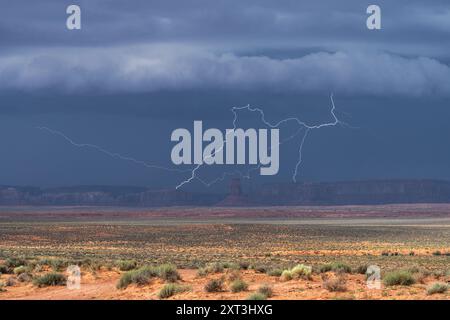 A powerful storm casting dark clouds unleashes a striking bolt of lightning over the iconic red desert landscape of Monument Valley Stock Photo