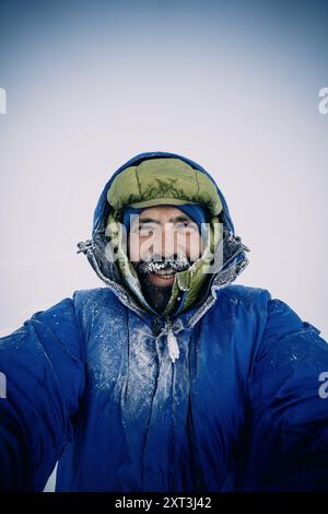 Self portrait of a man smiles warmly at the camera, his face partially covered in frost while wearing a jacket with hood, during a winter expedition i Stock Photo