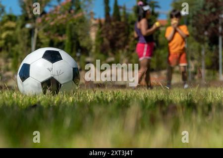 A soccer ball rests in the foreground on lush green grass with children playing games in the background of a vibrant park Stock Photo