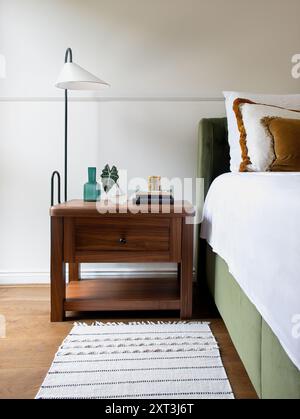 Modern bedroom interior showcasing a sleek wooden bedside table with a lamp, books, decorative vases, and green velvet headboard. Stock Photo