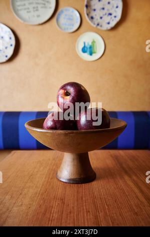 A tasteful display of shiny red apples in a rustic bowl on a wooden table, with decorative ceramic plates adorning the wall in the background. Stock Photo
