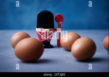 A whimsical soldier egg cup armed with a spoon stands guard among a formation of brown eggs on a soft blue background, creating a playful kitchen scen Stock Photo