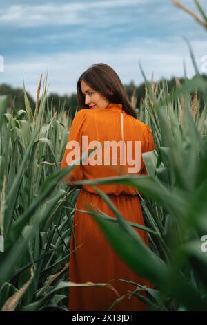 A brunette woman in an orange dress is seen looking away while standing amidst tall green cornstalks under a cloudy sky. Her gentle pose and thoughtfu Stock Photo