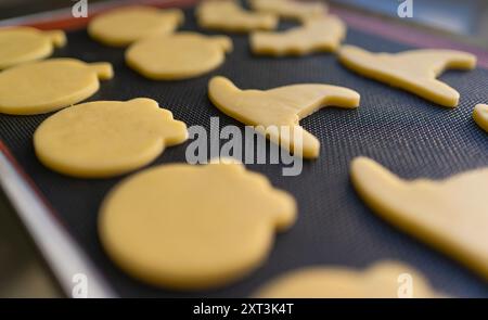 A close-up image of raw Halloween-themed cookies on a baking sheet The shapes include pumpkins and Witch hats, ready to be baked The focus is on the d Stock Photo