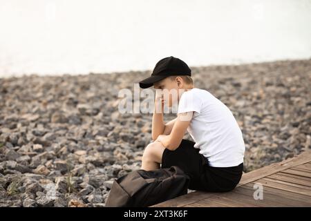 Thoughtful young boy sits alone on a wooden dock, gazing out over a rocky terrain. His expression is contemplative as he enjoys a quiet moment in the Stock Photo