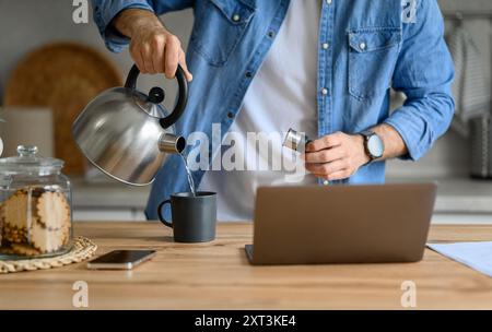 Midsection of businessman pouring hot water in coffee cup while using laptop on kitchen counter Stock Photo