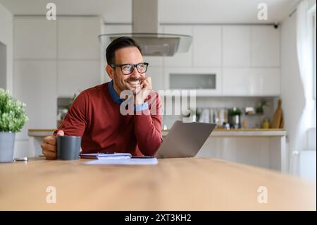 Thoughtful smiling businessman with hand on chin looking away while sitting at desk in home office Stock Photo