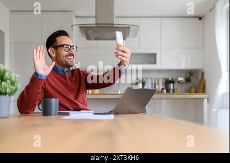 Happy businessman with laptop on desk waving hand while video calling over smart phone at home Stock Photo