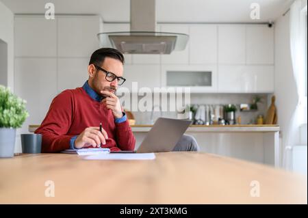 Businessman with hand on chin thinking ideas and writing on notepad while using laptop on desk Stock Photo