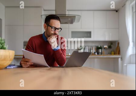 Focused businessman with hand on chin holding documents and looking at laptop in home office Stock Photo