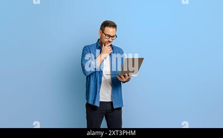 Serious handsome entrepreneur touching chin and doing research over laptop against blue background Stock Photo