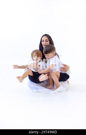 A young pregnant woman in a white dress with her cute sons. Adorable mom and her little kids are playing on the floor. White isolated background. A ha Stock Photo