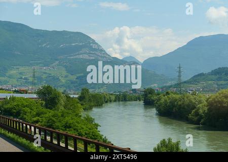 A scenic view of a road winding through a valley surrounded by majestic mountains, a river flowing alongside, and a clear blue sky with fluffy clouds. Stock Photo