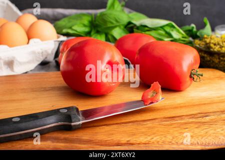 Cored Roma Tomato on a Wooden Cutting Board: Plum tomatoes and paring knife on a wood table with eggs, basil, and a dish of pesto on the background Stock Photo