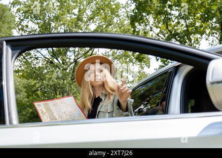 Blonde woman in hat staying next to car door checks the route on map to get to the destination. Young tourist explore local travel making candid real moments. True emotions expressions of getting away and refresh relax on open clean air Stock Photo